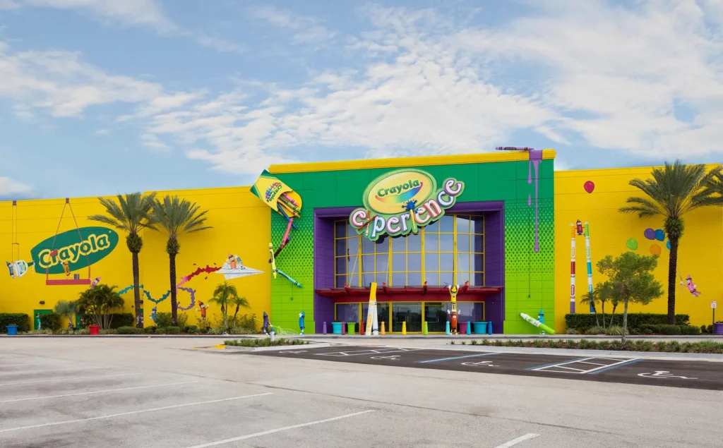 Fachada de Crayola Experience, un edificio gigante verde y amarillo con crayones gigantes colgando alrededor. Arriba un cielo celeste con una gran nube en el medio