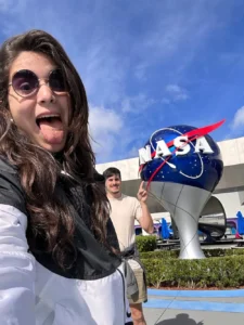 Juli al frente con una campera negra y blanca, sacando la lengua, atrás se ve a su novio señalando la bola gigante representativa de la NASA. Azul brillante, con el nombre "NASA" en blanco y un rayo rojo que lo cruza.