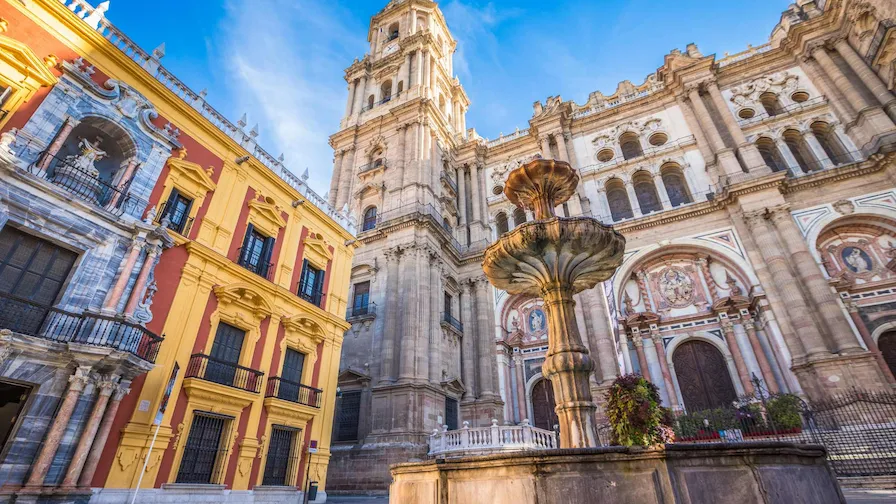 Fachada de la Catedral de Málaga. A la izquierda un edificio luce los colores de España rojo y amarillo. Frente a los edificios una fuente con agua de estilo medieval.