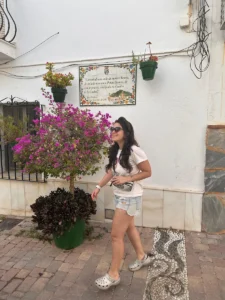 Juli camina por una de las calles, en la pared detrás se ve unos azulejos con un poema. Estepona está lleno de poemas en sus paredes. Al lado de ella un árbol pequeño con flores rosas